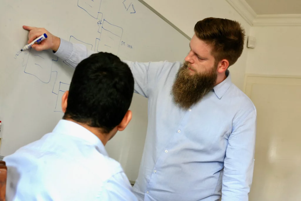 A man in a light blue shirt is pointing at a whiteboard with a marker, explaining a diagram to another person.