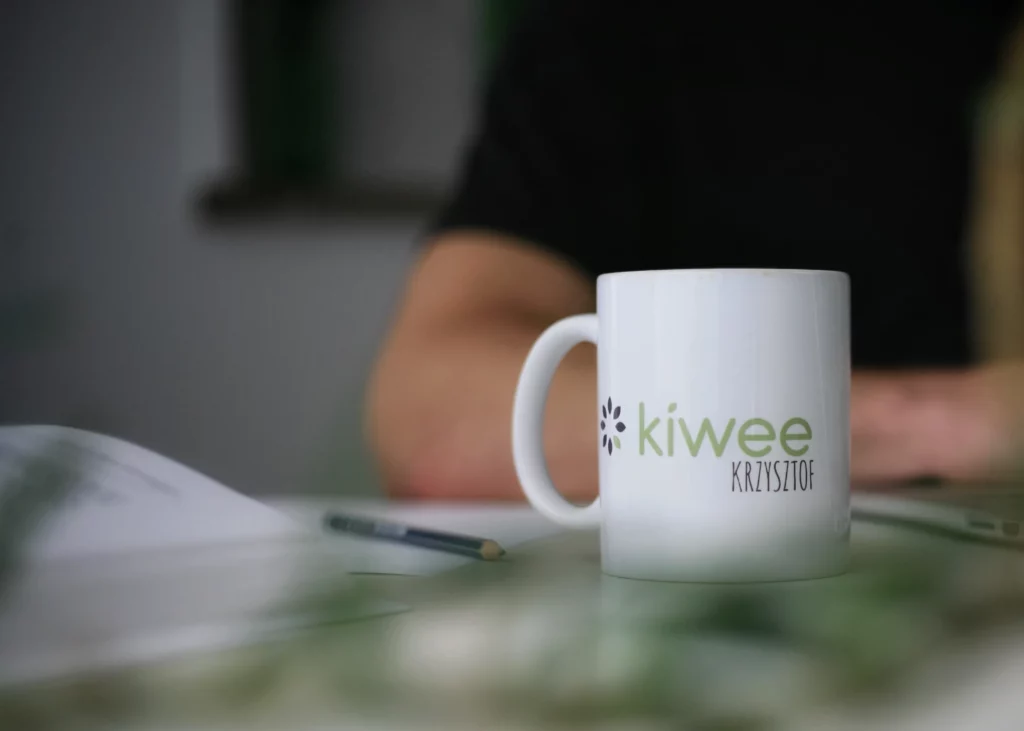 A white mug with the word "Kiwee" printed on it is placed on a table. A person's blurred arm is seen in the background, along with a pencil and some papers.