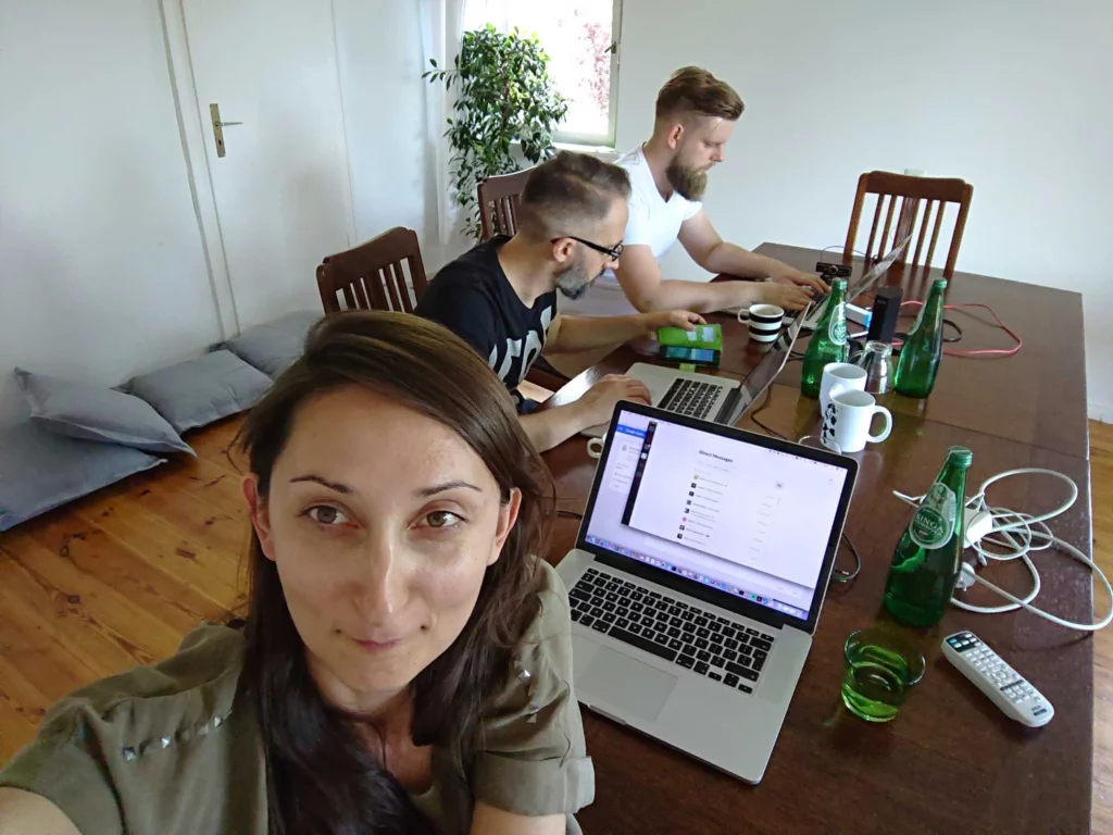 Three people are working at a wooden table with laptops. A woman in the foreground is taking a selfie, while two men are focused on their screens.