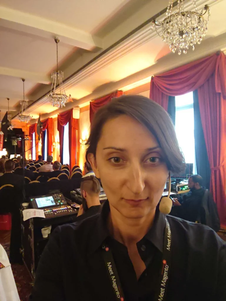 A person with short brown hair takes a selfie during a conference in a well-lit room with chandeliers, red curtains, and rows of black chairs. The person is wearing a black shirt.