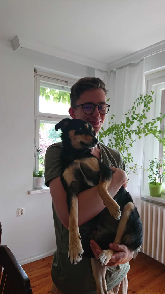 Eine lächelnde Person mit Brille hält in einem der Büroräume einen schwarzen Hund im Arm.
