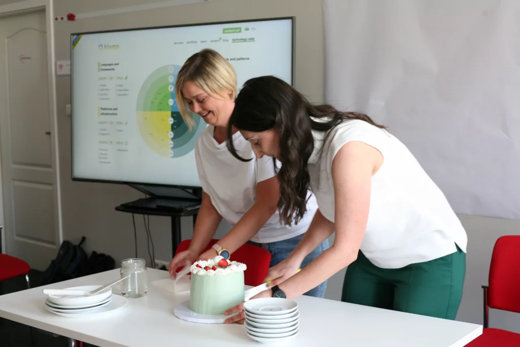 Two women preparing to cut a cake in an office setting. A presentation slide is visible on a screen in the background.