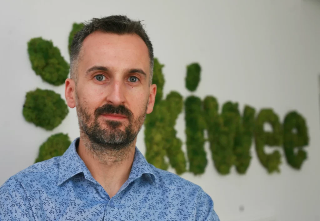 A man with a beard and short hair wearing a blue patterned shirt is standing in front of a green moss Kiwee logo.