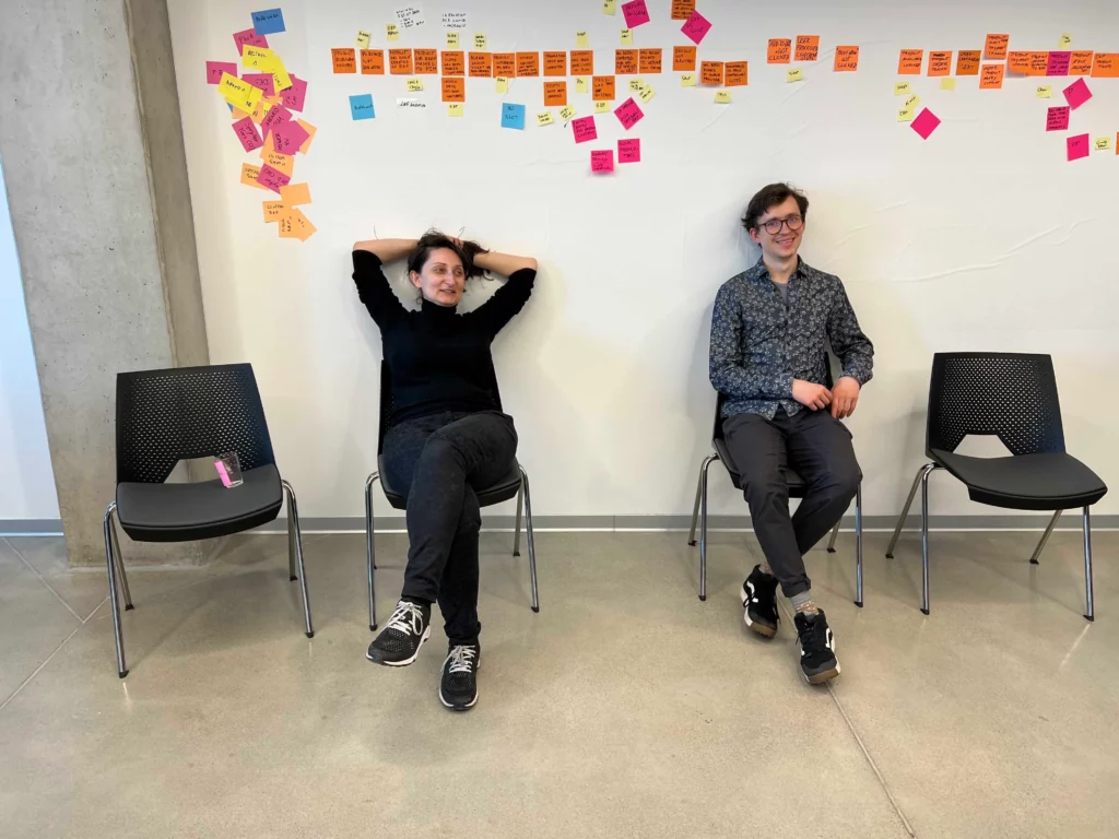 Two people sitting on chairs in a room with a white wall covered in colorful sticky notes.