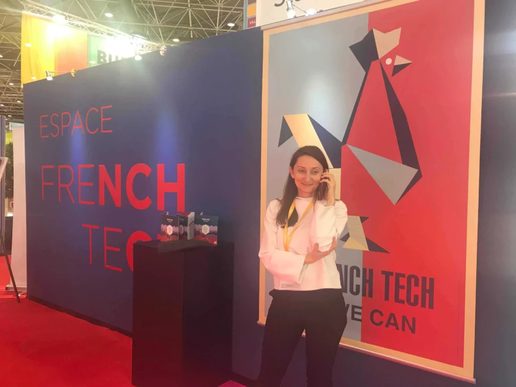 A woman stands smiling and talking on the phone in front of a "FRENCH TECH" conference sign.