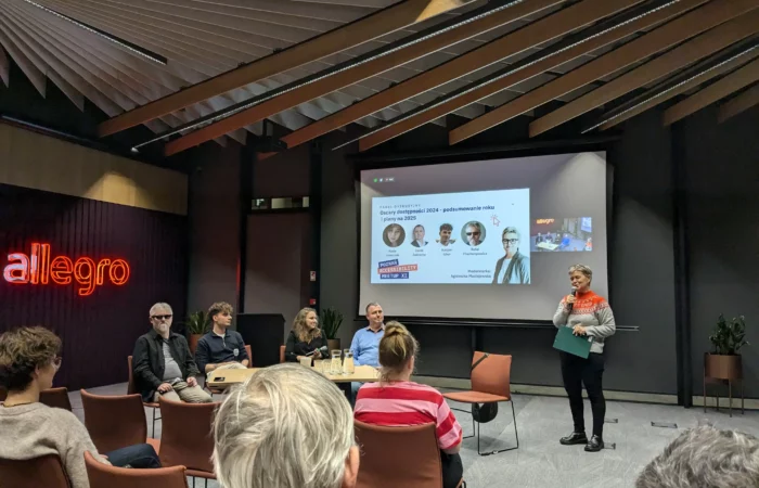 A panel discussion with five people seated at a table and one person standing, presenting a slideshow in a conference room.