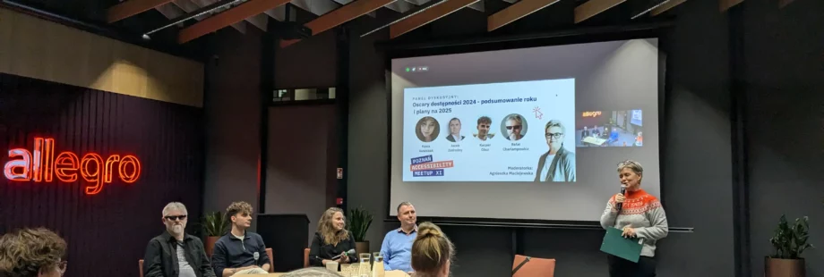 A panel discussion with five people seated at a table and one person standing, presenting a slideshow in a conference room.