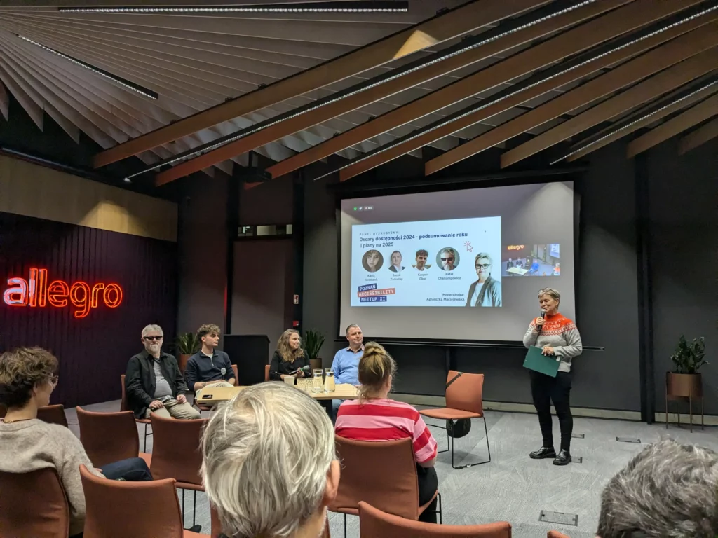 Eine Podiumsdiskussion mit fünf Personen, die an einem Tisch sitzen, und einer Person, die steht und eine Diashow in einem Konferenzraum präsentiert.