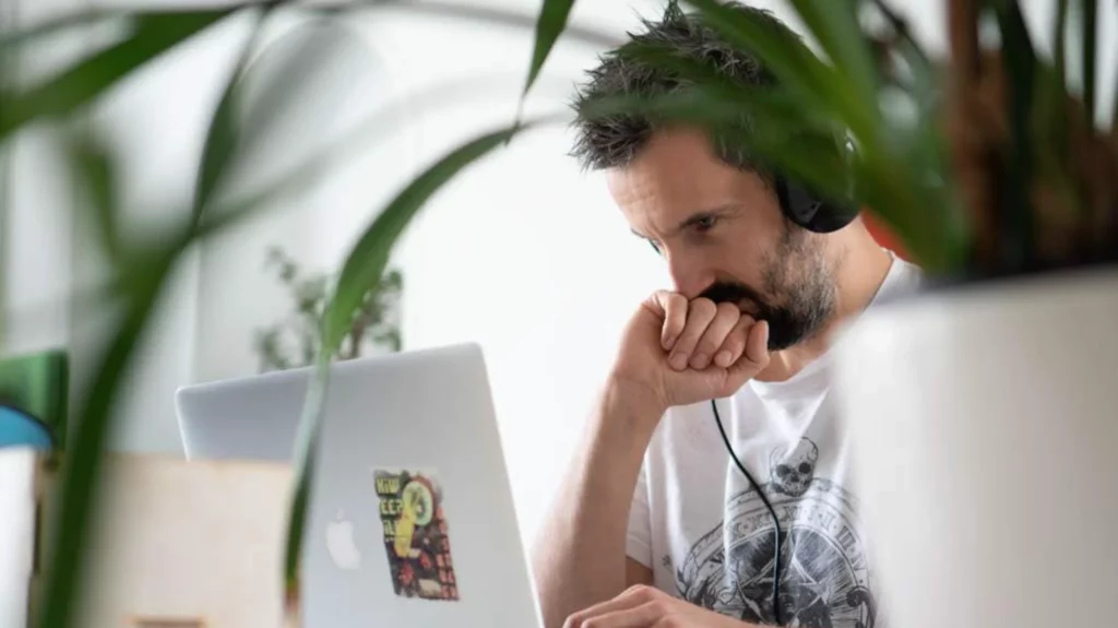 Man wearing headphones and working on a laptop in an office surrounded by plants.